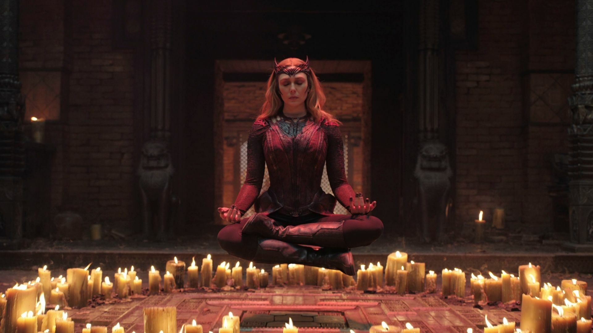 A woman dressed in red floats, seated, above a circle of lit candles.