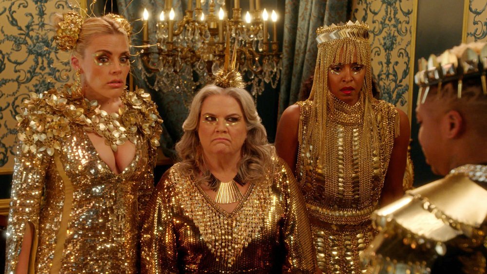 Three women in elaborate gold outfits, accessories, and makeup, on set but not actively filming a music video; still from "Girls5eva."