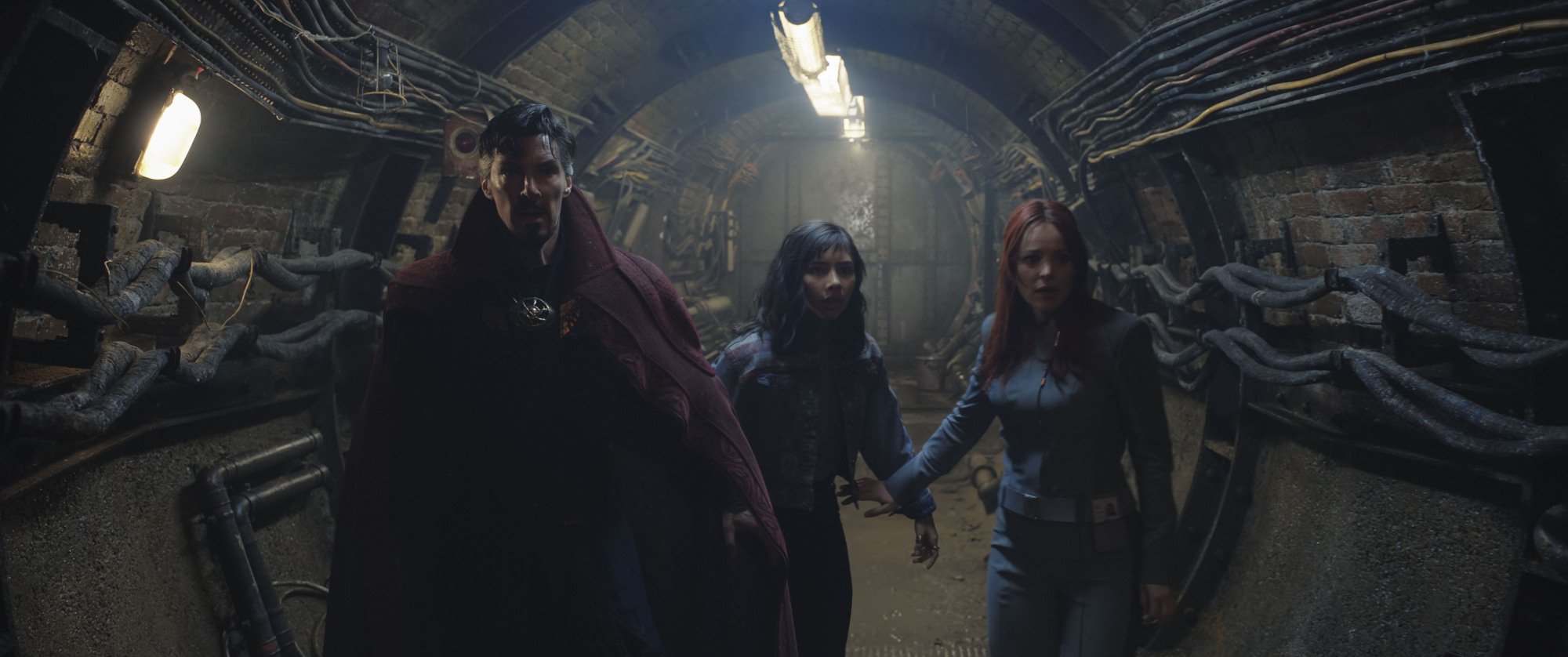 A man, a young girl, and a woman walk through an underground tunnel lined with pipes.