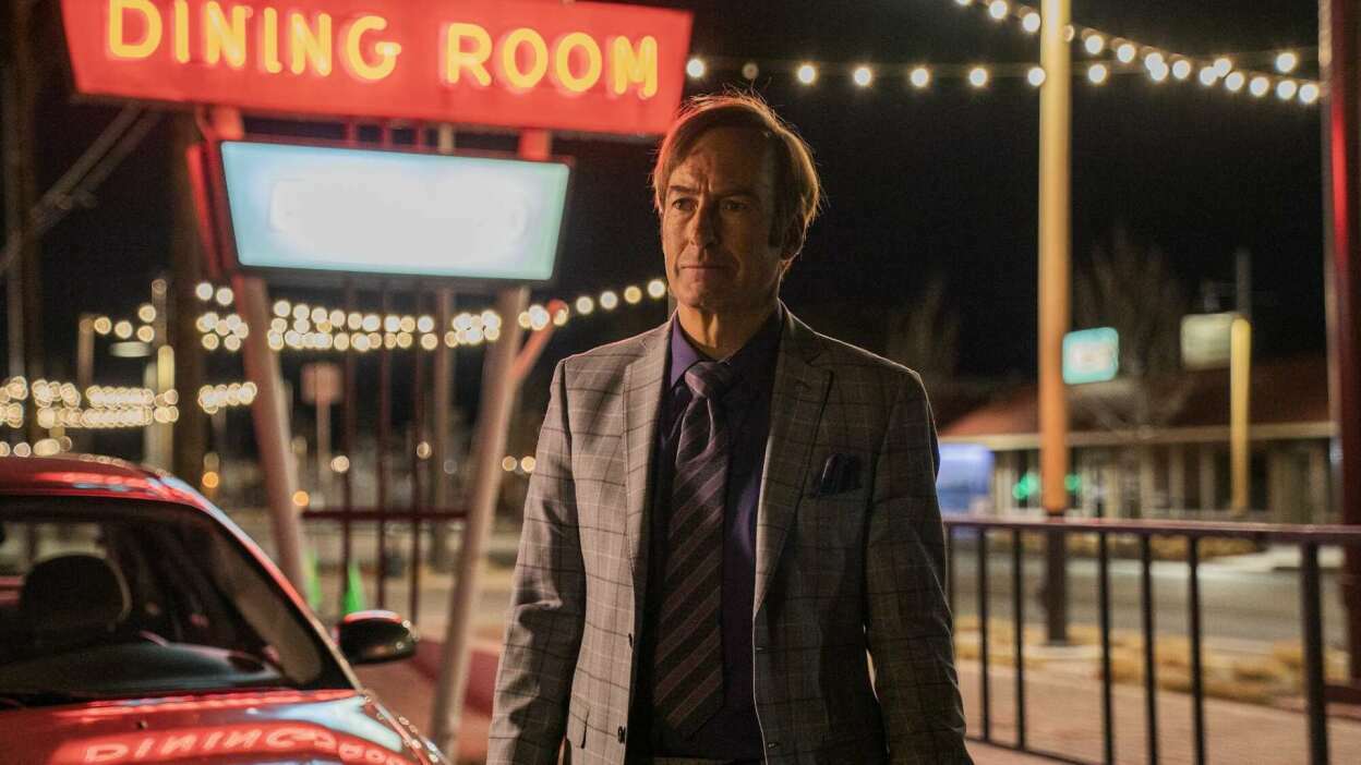 A man in a suit standing next to a car and a neon sign that says "dining room."