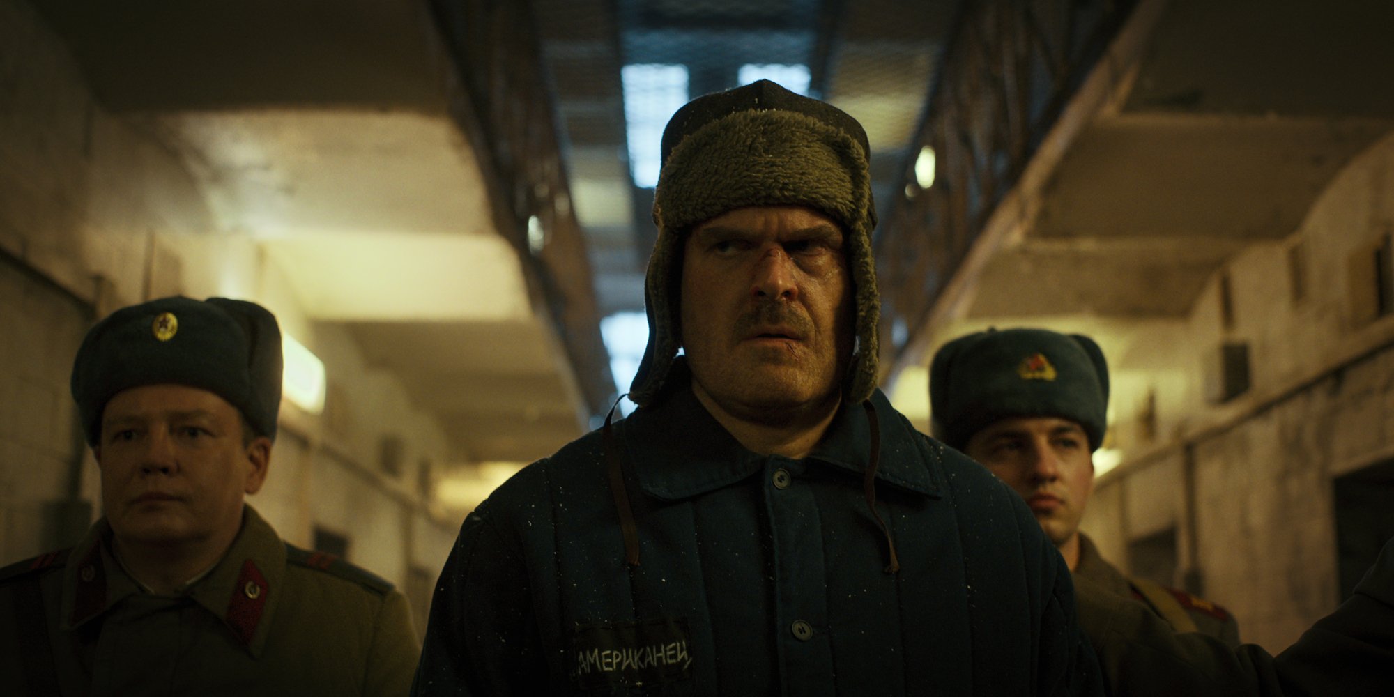 Three men in heavy coats and furry hats walk through a prison block.