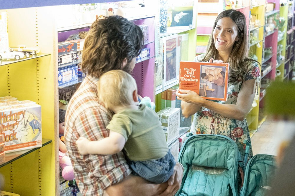 A man (Milo Ventimiglia as Jack) holding a baby in a store while a woman (Mandy Moore as Rebecca) holds up a Pin The Tail on the Donkey game.
