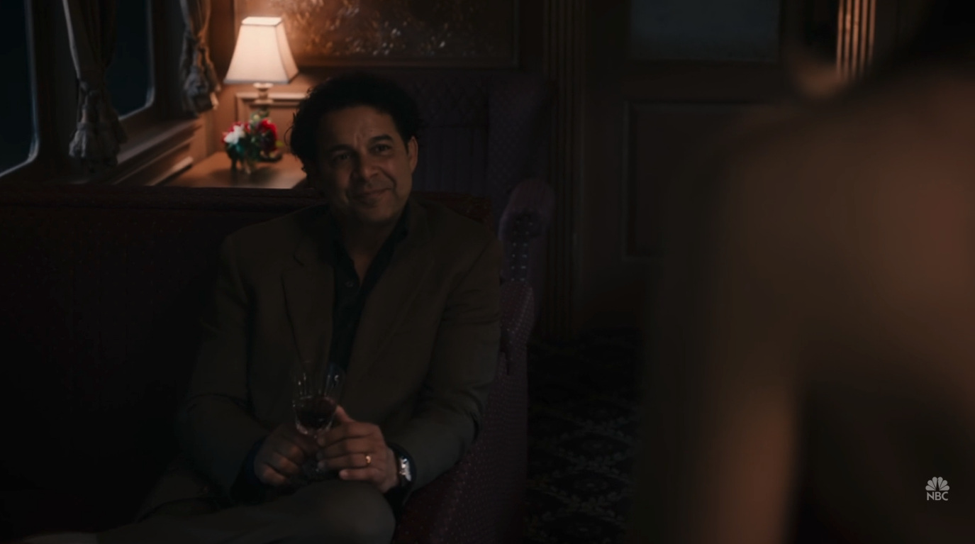 A man (Jon Huertas) sitting on a train holding a glass of wine.