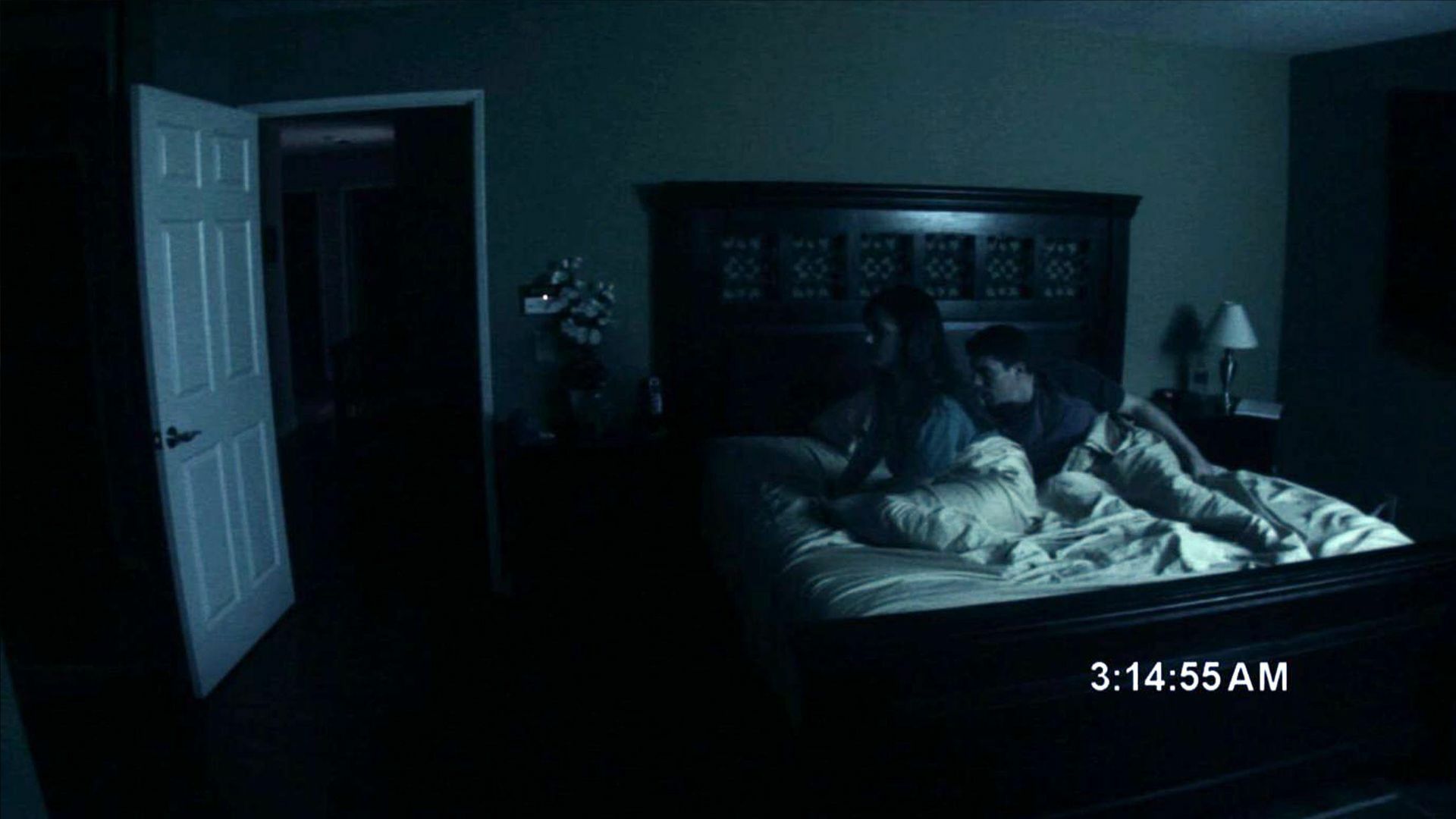 In a dark room, a couple lies in bed while staring, alarmed, at an open door. 