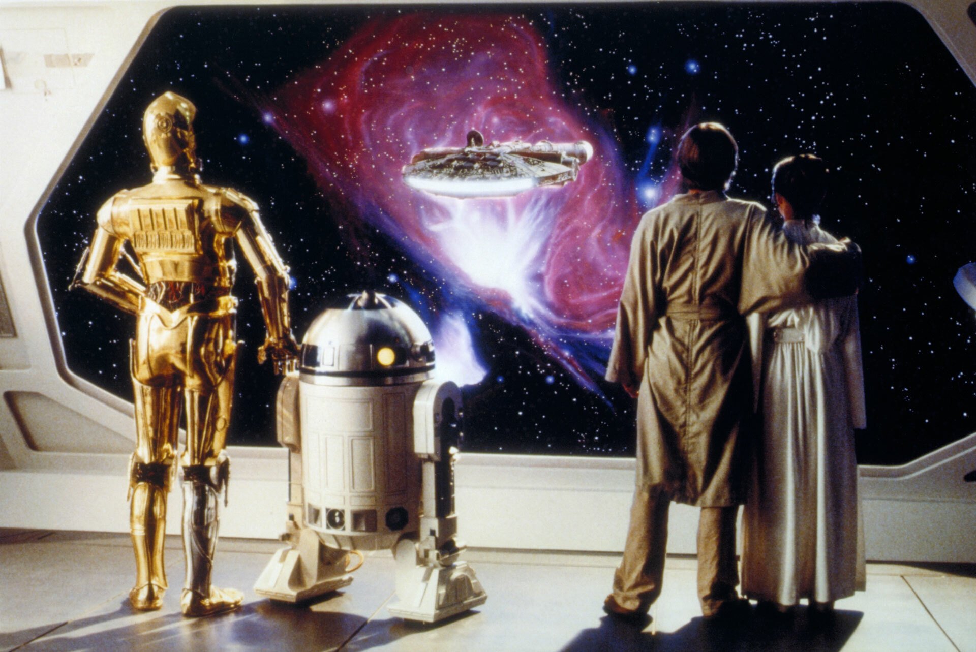 A still from the movie "Star Wars: The Empire Strikes Back." C-3P0, R2-D2, Luke Skywalker, and Princess Leia stand facing a window into space where they see the Millennium Falcon in flight.