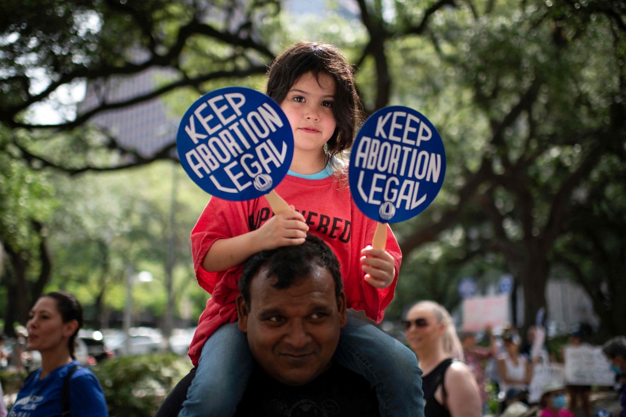 A young child sits on the shoulders of an adult. They are holding two small signs that read "Keep abortion legal". 