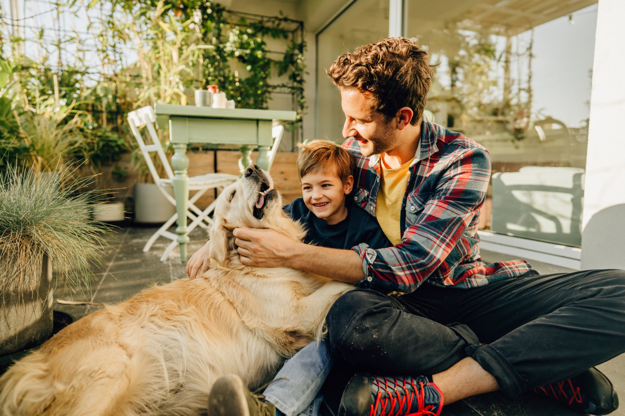 Image of father and young boy sitting on the ground petting a golden retriever who is happy and relaxed