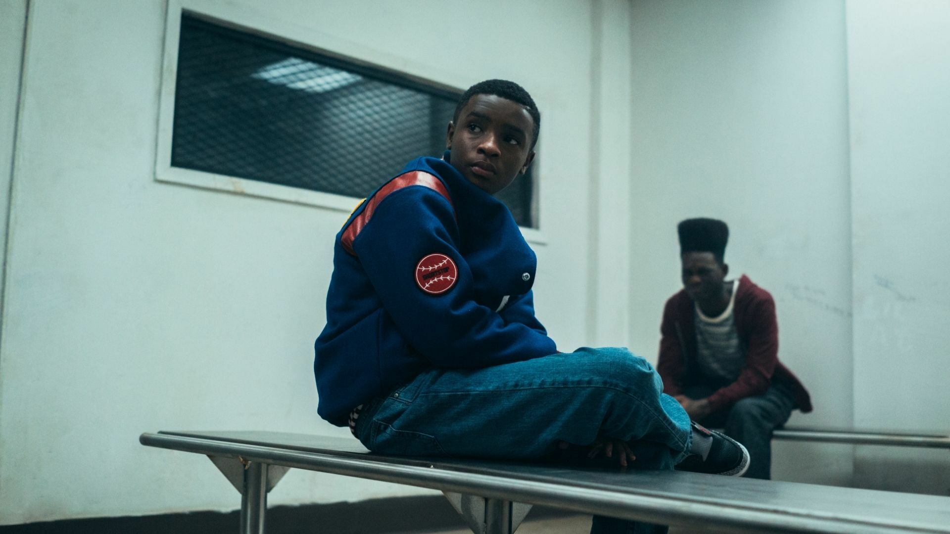 A young Black boy in a baseball jacket sits on a bench with another Black boy in the background, from "When They See Us"