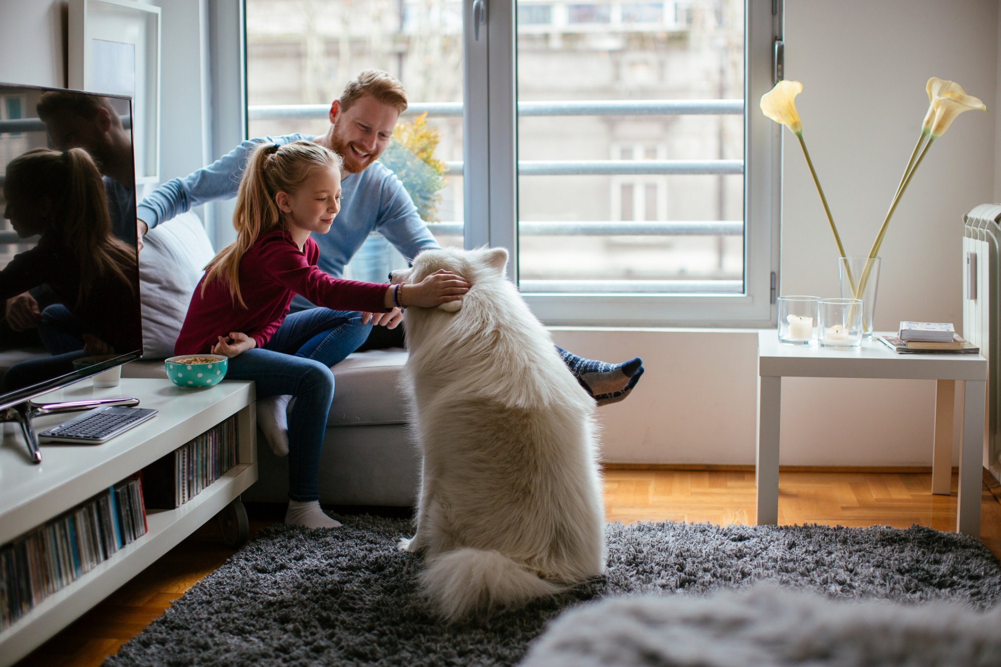 Image of father and young girl sitting on a chair petting a dog who is sitting and faced towards them.