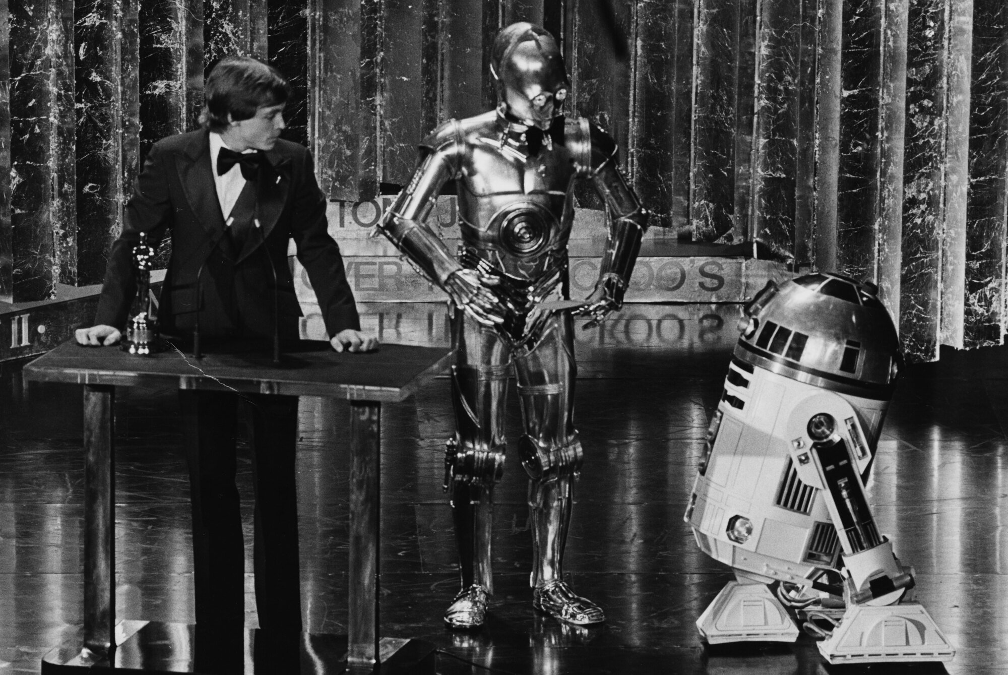 Mark Hamill stands at a podium on a stage. He's wearing a tuxedo, and C-3P0 and R2-D2 are right next to him.
