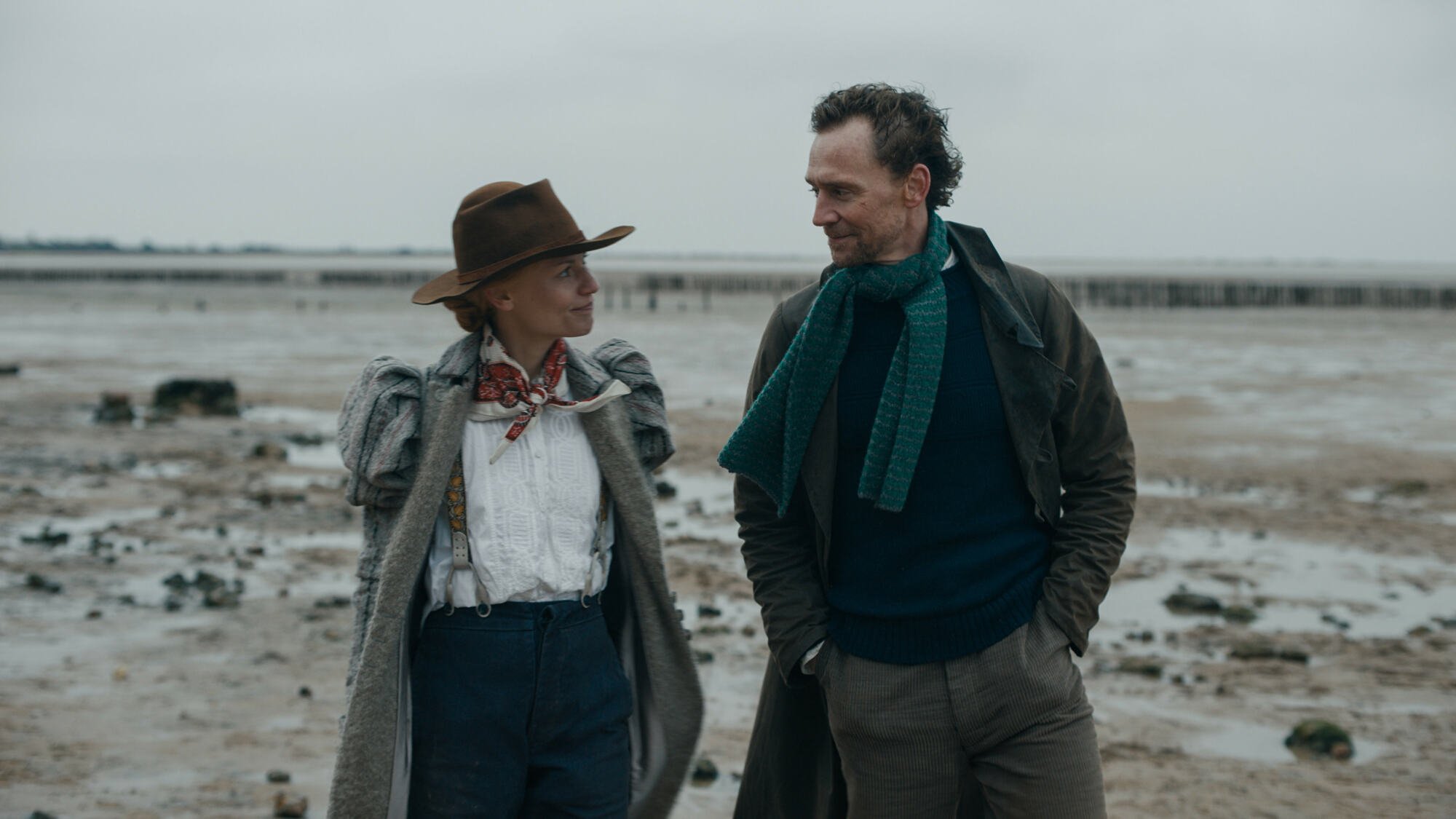 A woman and a man wearing old-fashioned warm clothes on a grey beach.