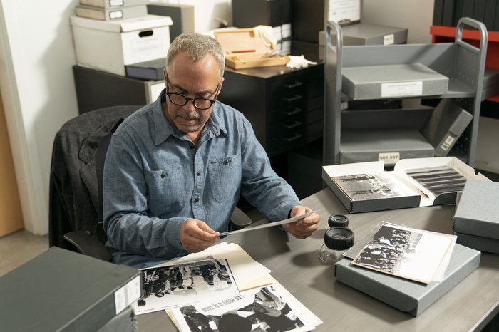 A man sits at a desk looking through black and white photographs.