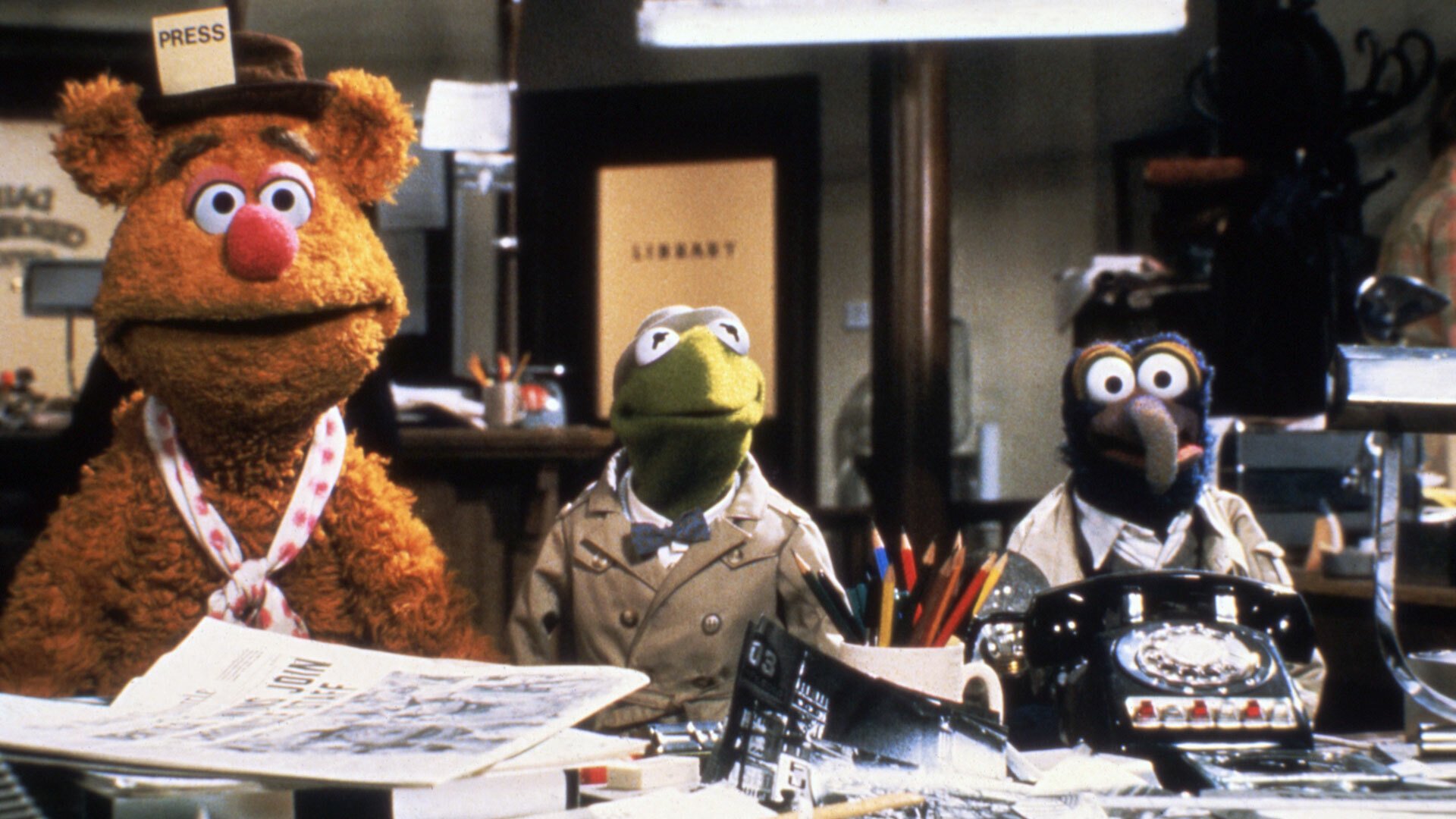 A still from "The Great Muppet Caper." Fozzie Bear, Kermit, and Gonzo stand side by side in front of a desk, in what appears to be a cluttered newsroom.