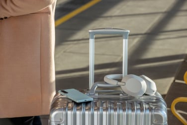 silver Sony WH-1000XM5 Headphones wrapped around the handle of a silver suitcase next to a phone and someone wearing a beige coat