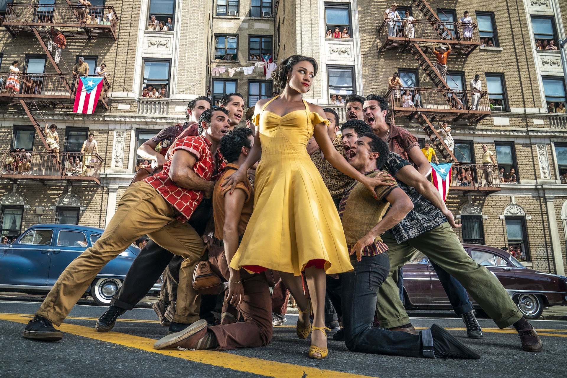 Ariana Debose as Anita in "West Side Story," wearing a yellow dress and surrounded by ensemble members.