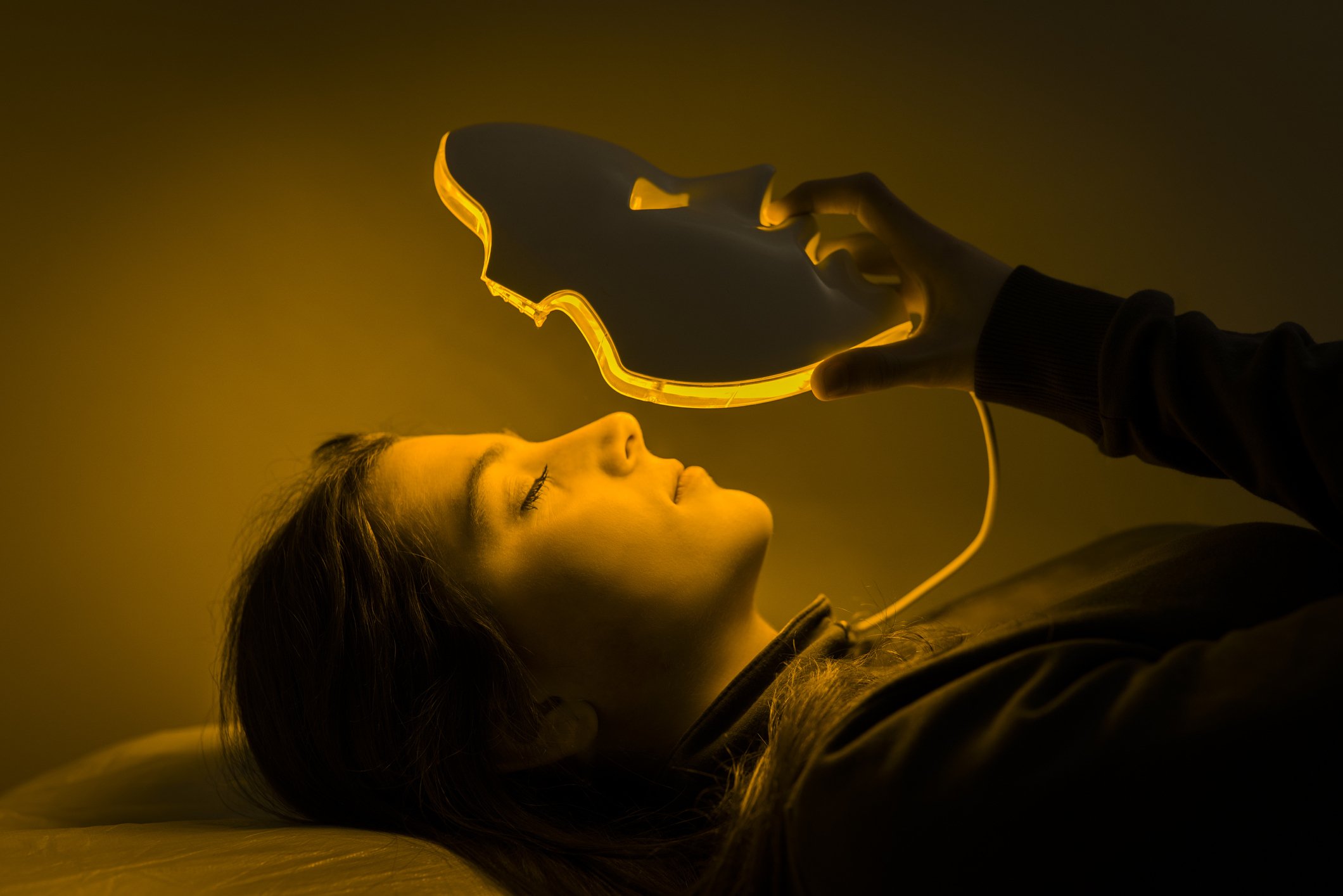 Woman laying down is putting on a face mask with eye, nose, and mouth cutouts. The mask is lit with yellow colored lights from within. 