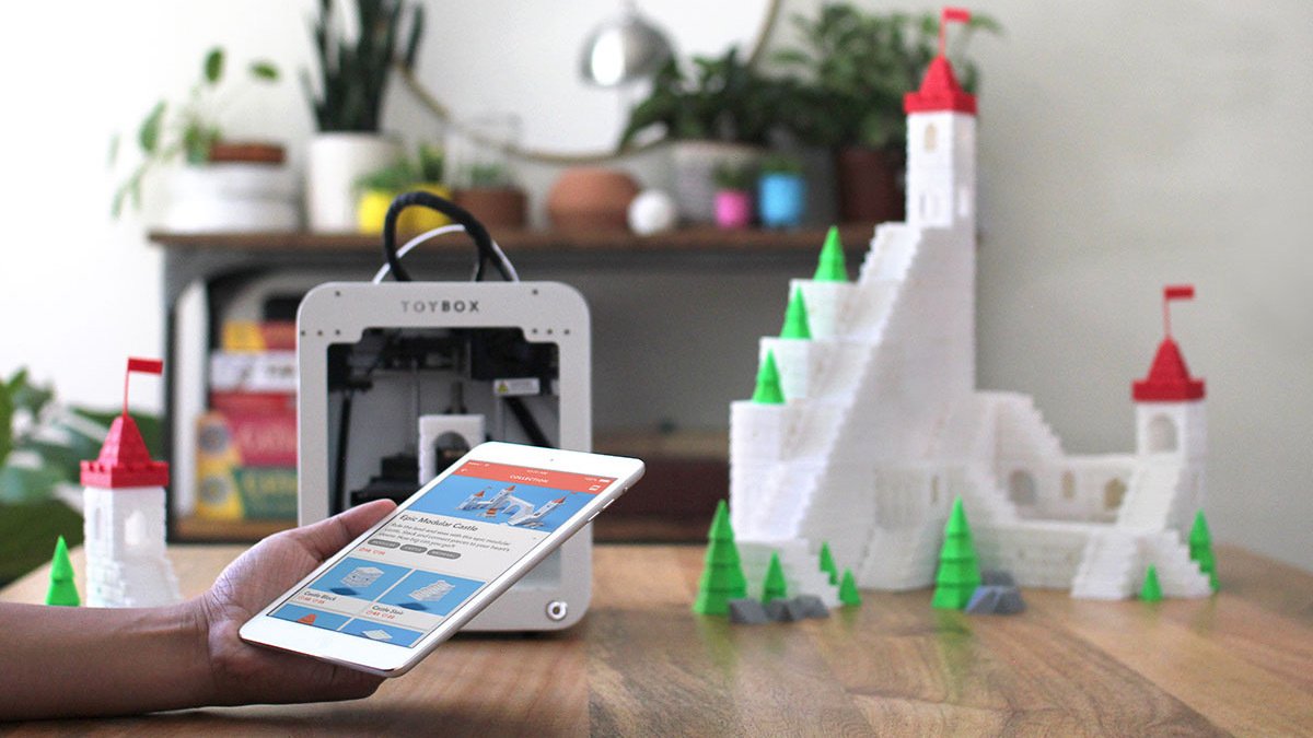 Hand holding ipad with castle design with 3d-printed castle next to 3d printer