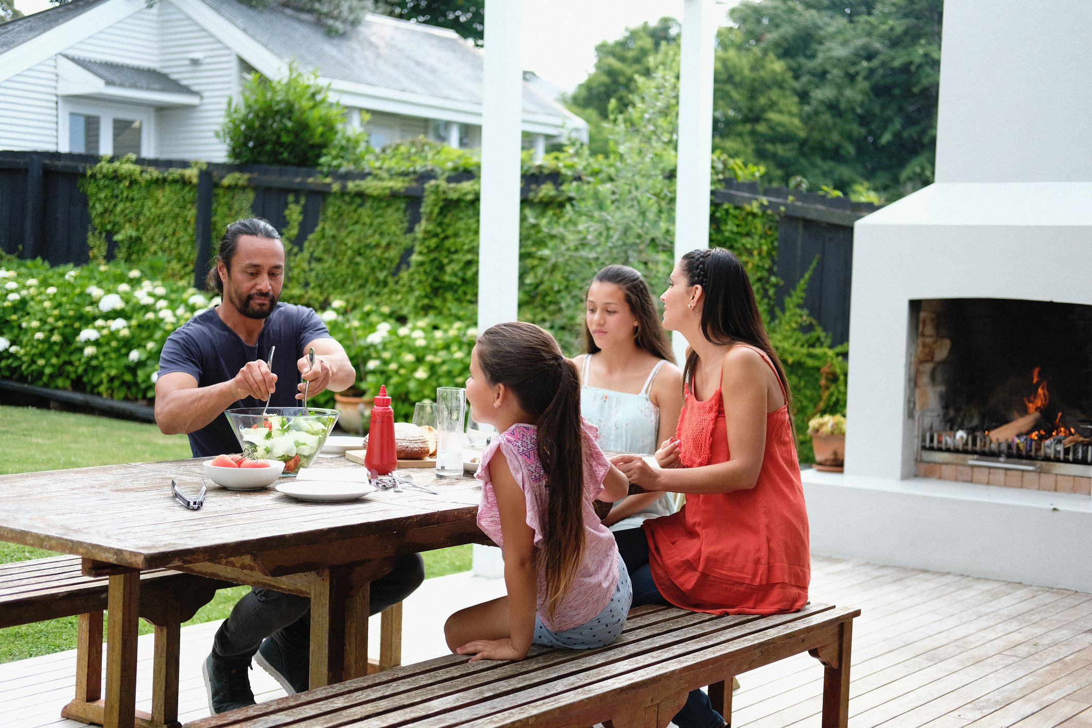 A family of four - mother, father and two young daughters - are enjoying a summer lunch on the outdoor backyard patio of their home
