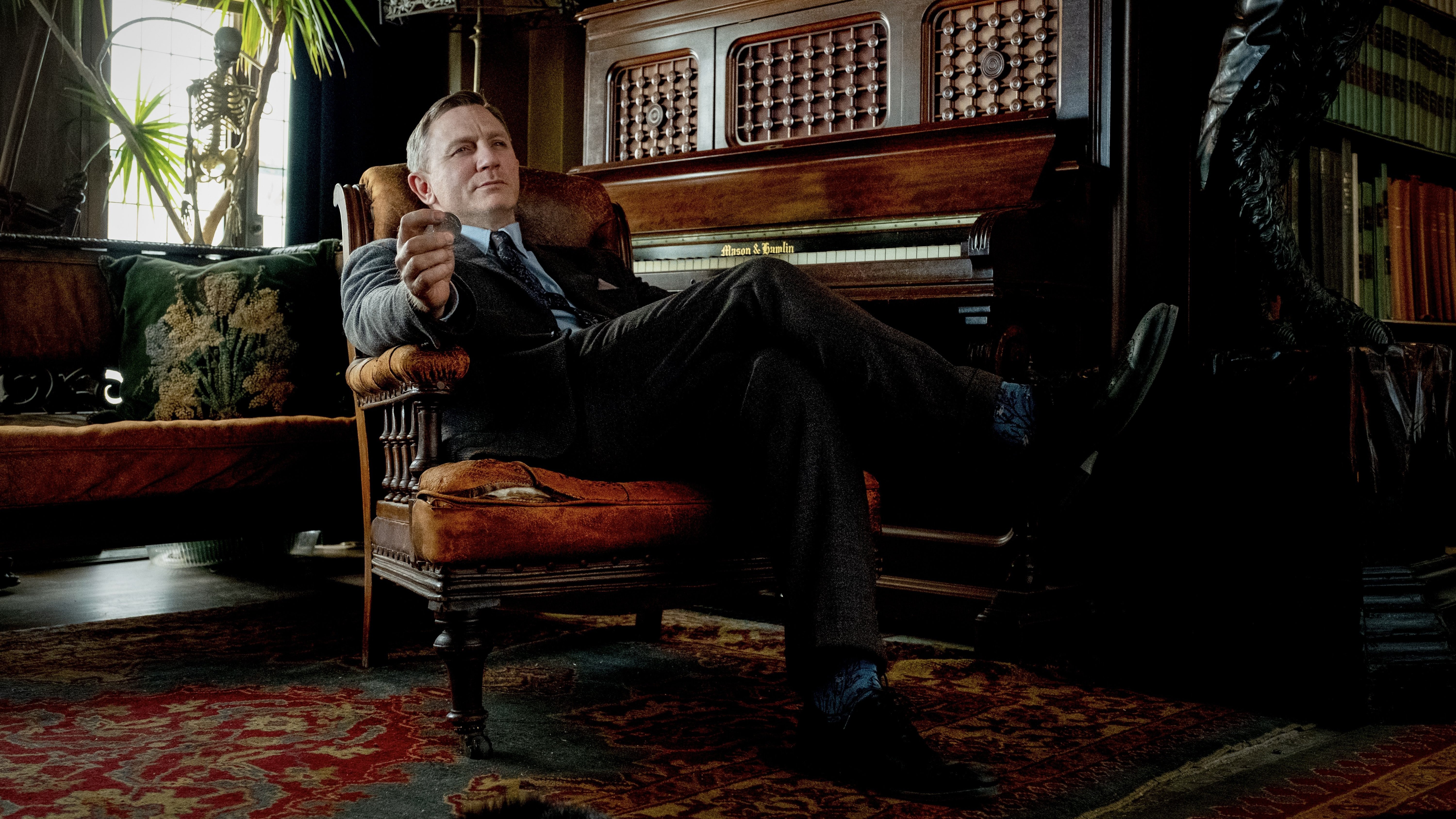 A man in a grey suit sits back in a fancy armchair beside a piano.