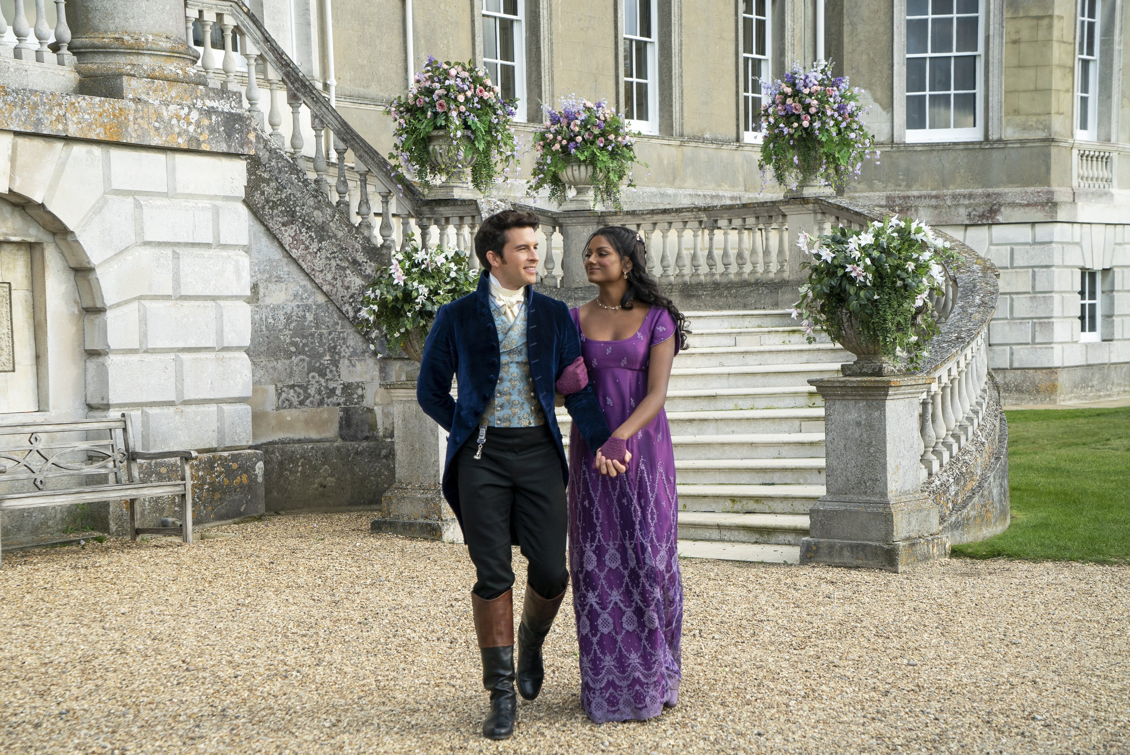 Two people in Regency costume hold hands outside a historic English house. 