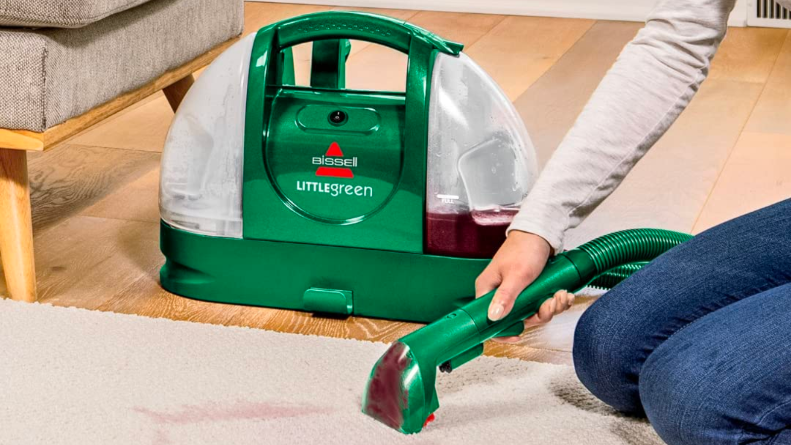 a woman in blue jeans kneels on a white carpet while removing a wine stain from it using the bissell little green cleaner