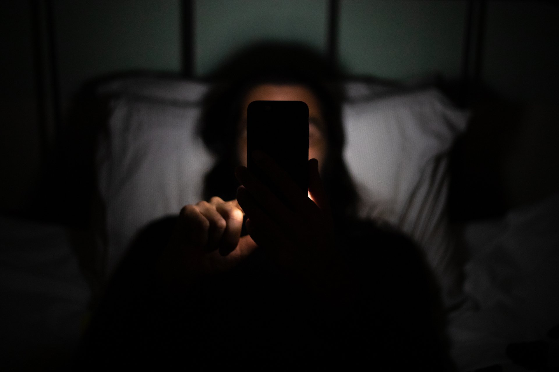 A person lays in bed in the dark, scrolling on a phone. Their face is obscured by the device. 