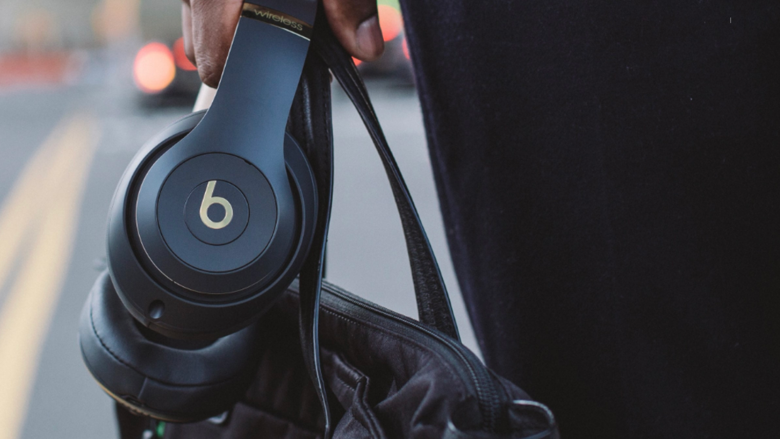 a close-up shot of someone carrying a pair of black beats studio3 headphones down the street. they're holding the straps of a black leather bag in the same hand