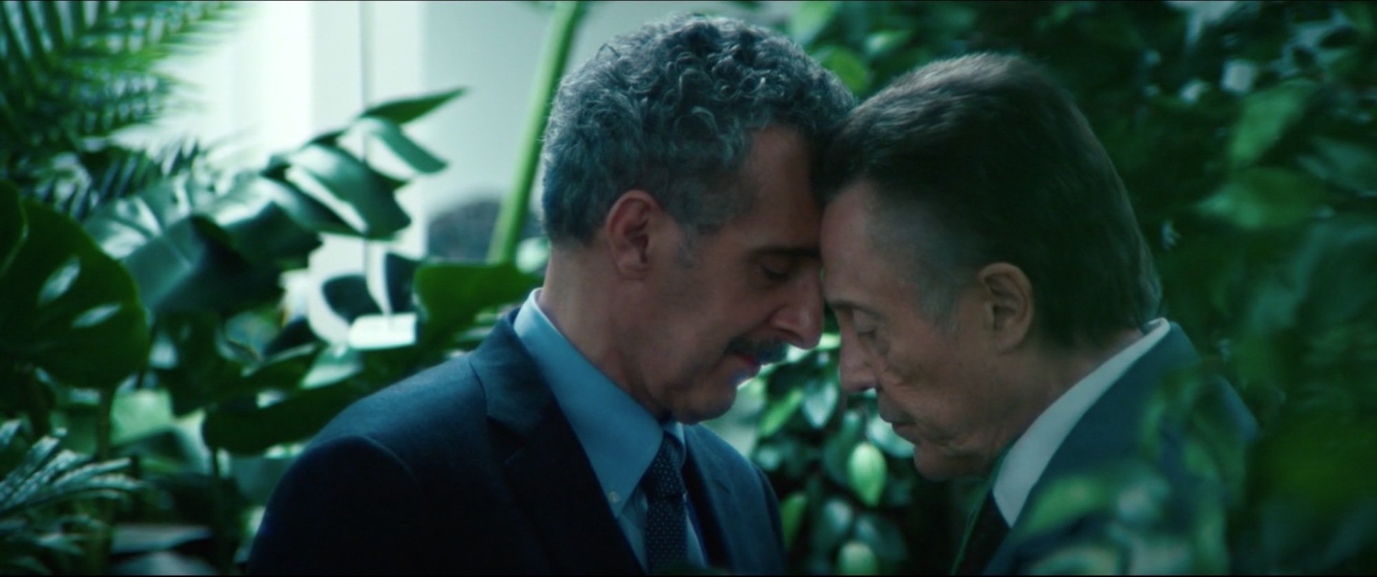Two men standing forehead to forehead in a room surrounded by plants.