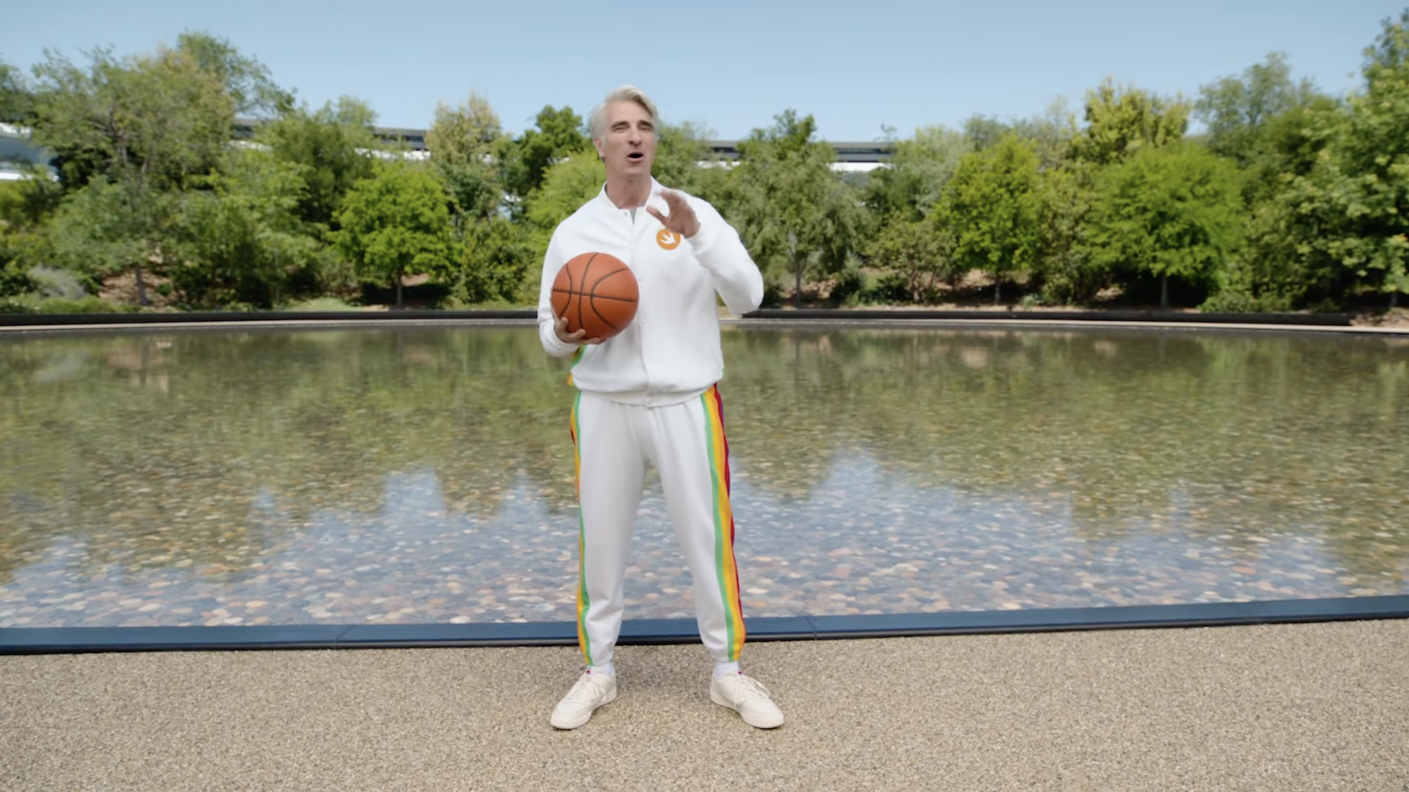 Craig Federighi holds a basketball in his palm and wears a white tracksuit with multi-colored stripes down the side.