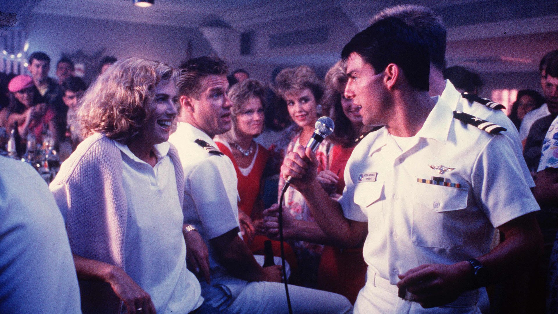 A man in navy uniform sings to a woman in a bar surrounded by people.