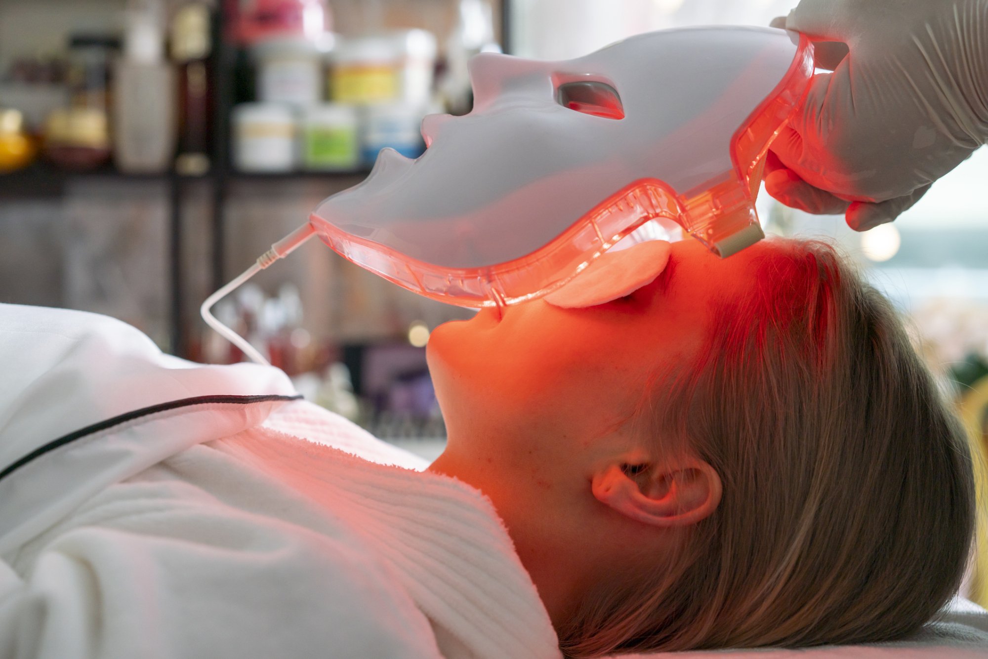Woman laying down with eye protection on, a white full face mask with red glowing lights is lowered over her face.