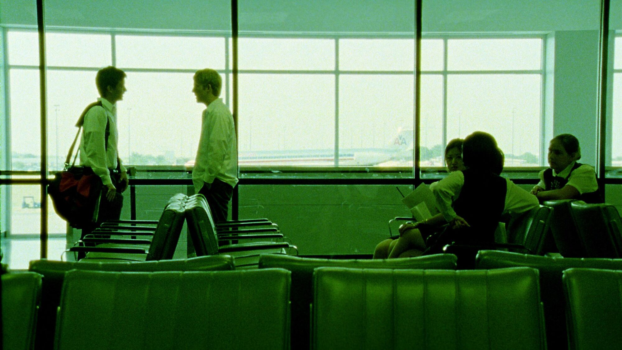 Two men stand on the left hand side of an airport scene, bathed in green light.