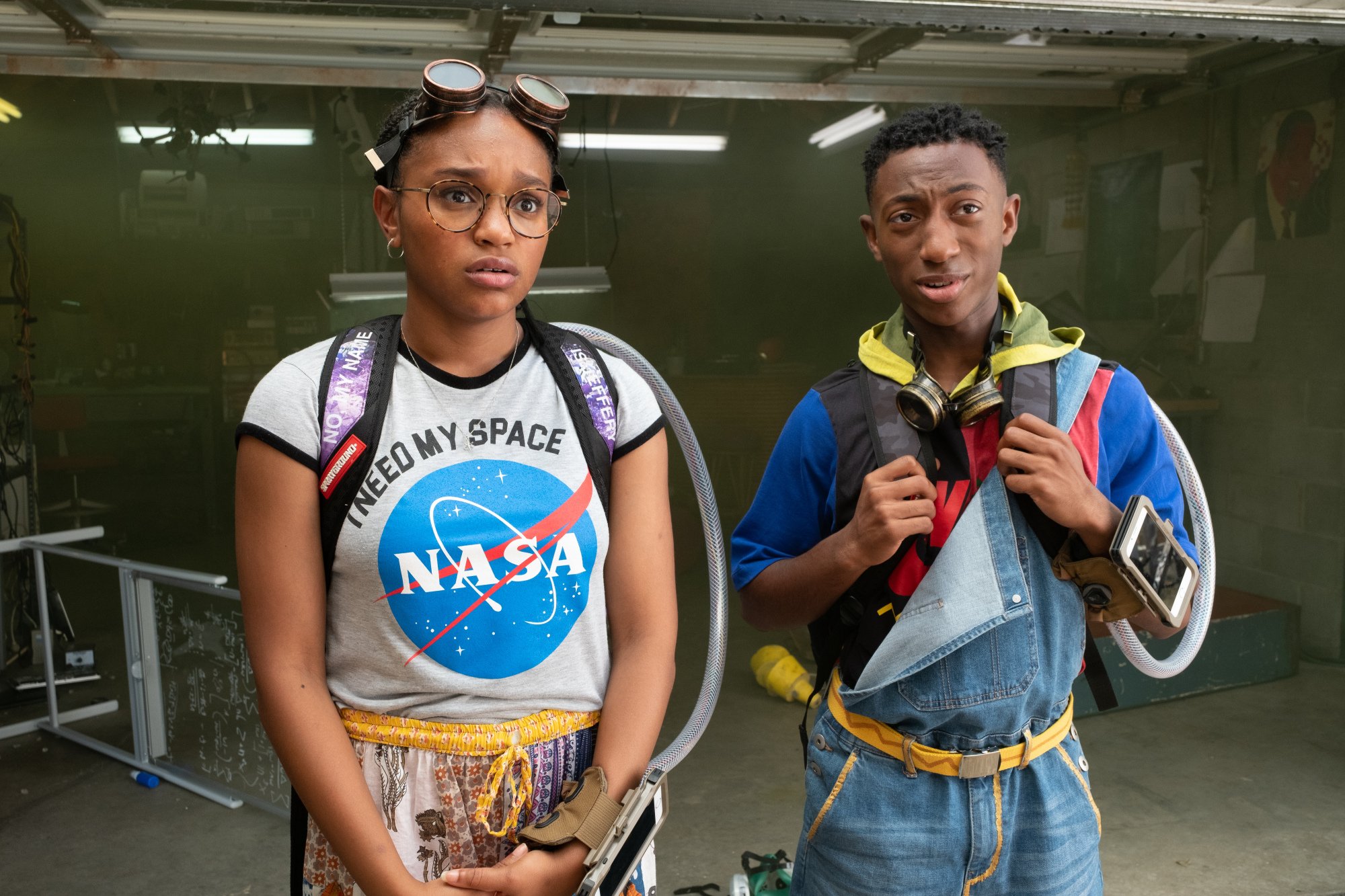 Two young people in backpacks, one wearing a NASA t-shirt, stand in a garage.
