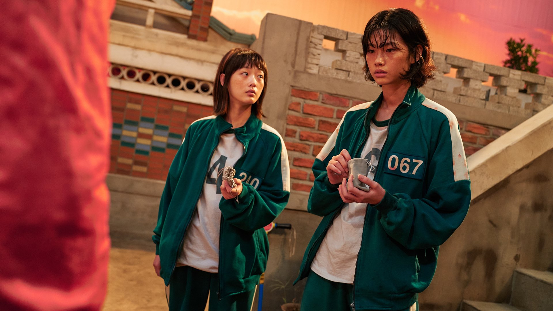 Two people in green jumpsuits stand holding small pouches a laneway set.