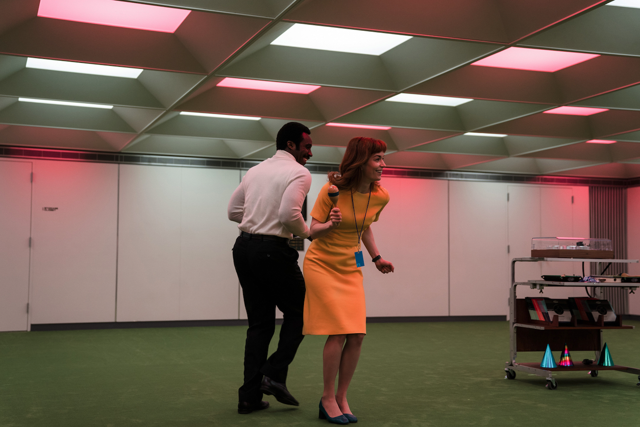 A man and a woman dancing in an office under white and pink flashing lights.