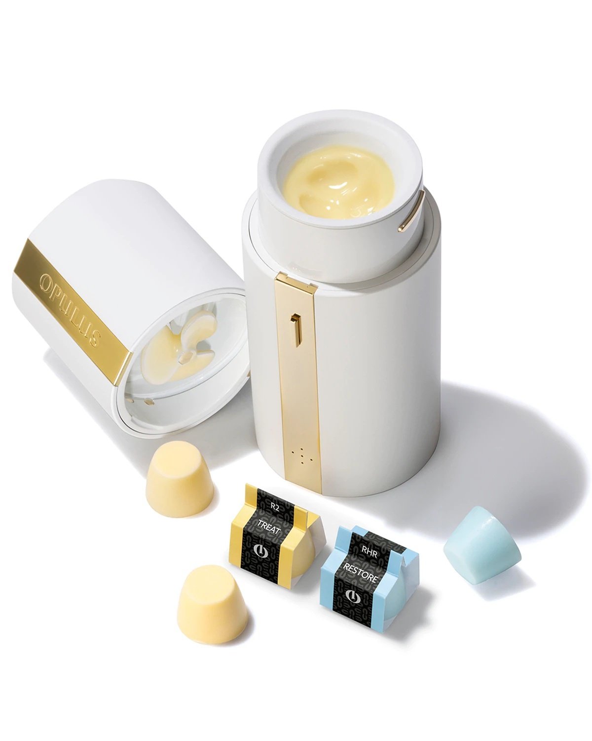 White cylindrical device with gold stripe in the middle. The cap is off to reveal a light yellow lotion-like skincare treatment inside. Small pods with more treatment sit next to it. 