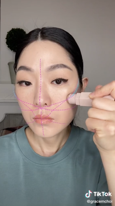 Asian woman with dark hair looks at camera and applies contour to cheek with a filter overlaid.