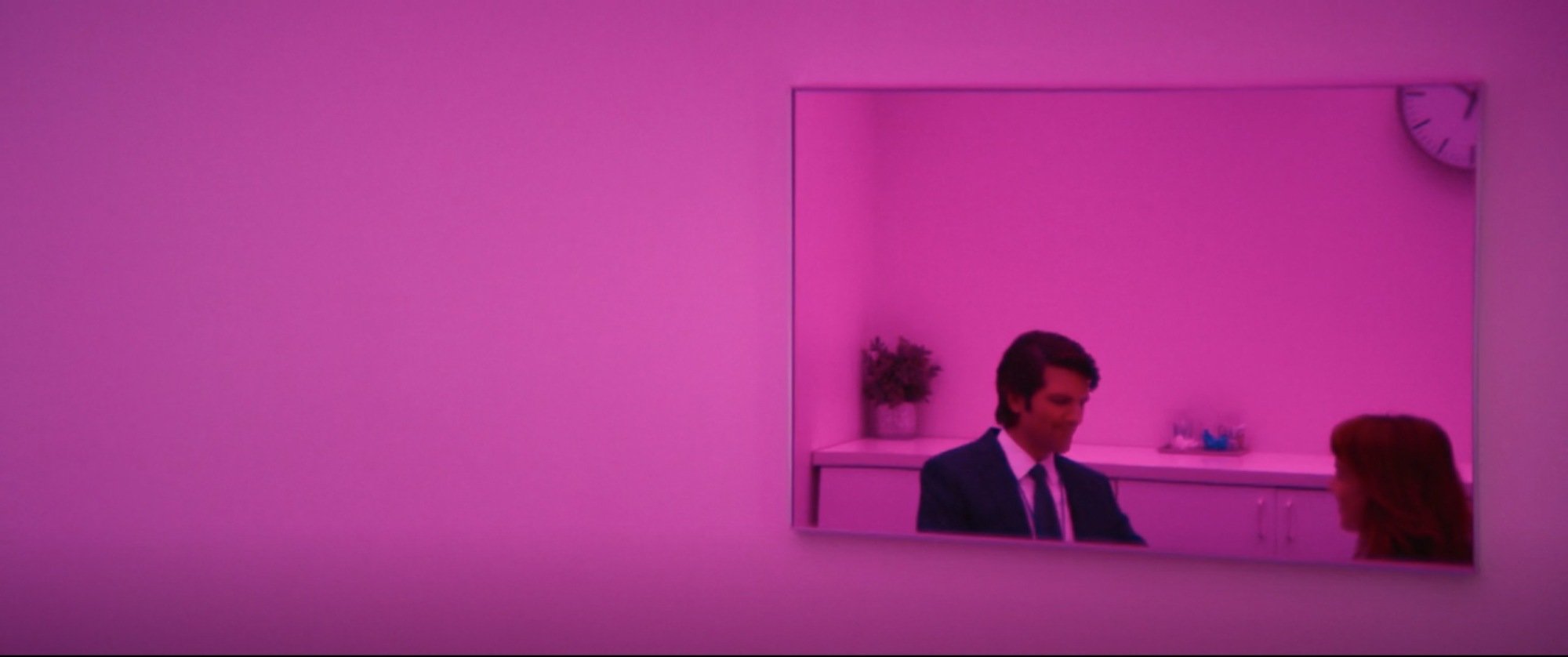 The reflection of man and a woman seen in a mirror in a room with pink lighting.