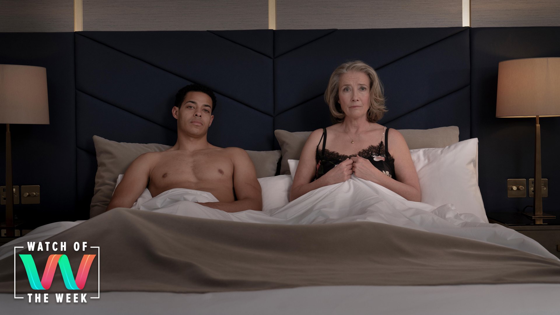 A man and woman in their underwear sit in a hotel bed with the covers pulled up, looking at the camera.