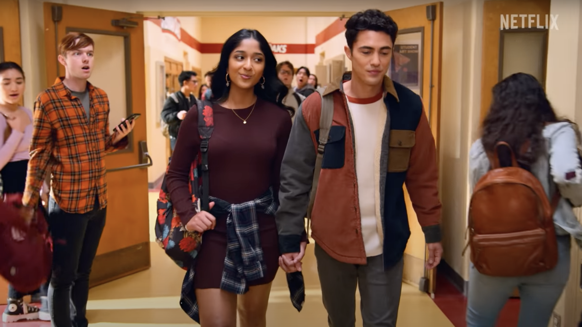 A high school couple walk through a hallway while people stare.