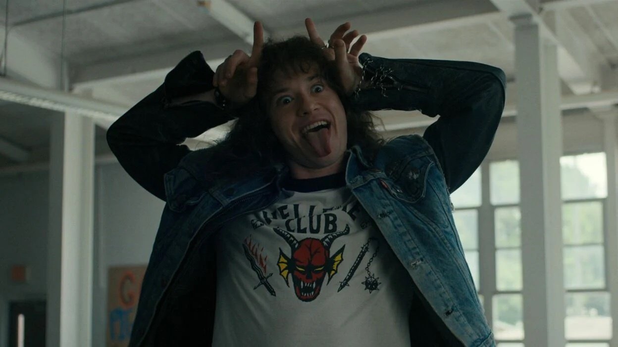 A young man in a shirt with a devil on it makes devil horns above his head.