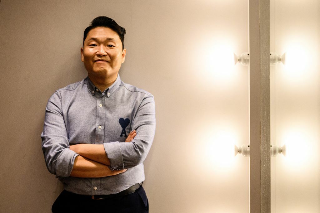 Psy stands against a wall in a blue button down shirt
