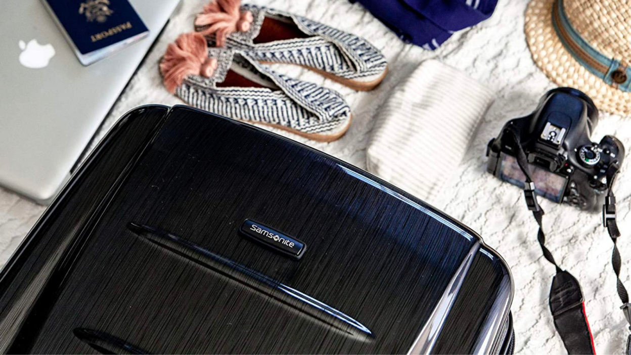a samsonite suitcase laid out on a white quilt next to a wicker hat, a camera, a folded white shirt, tassled shoes, and a macbook with a passport on it