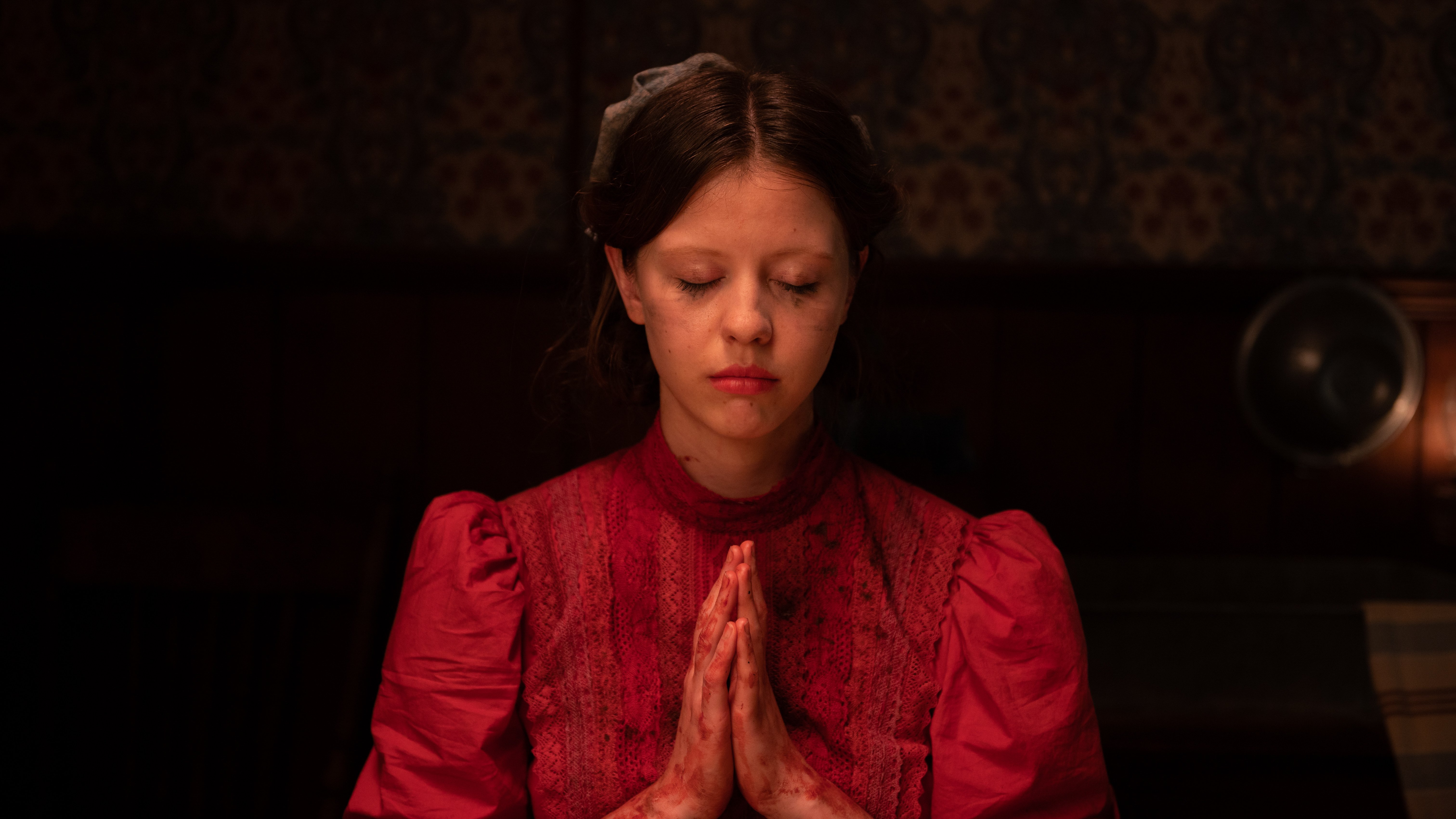 A girl in a blood-stained red dress prays with her eyes closed.