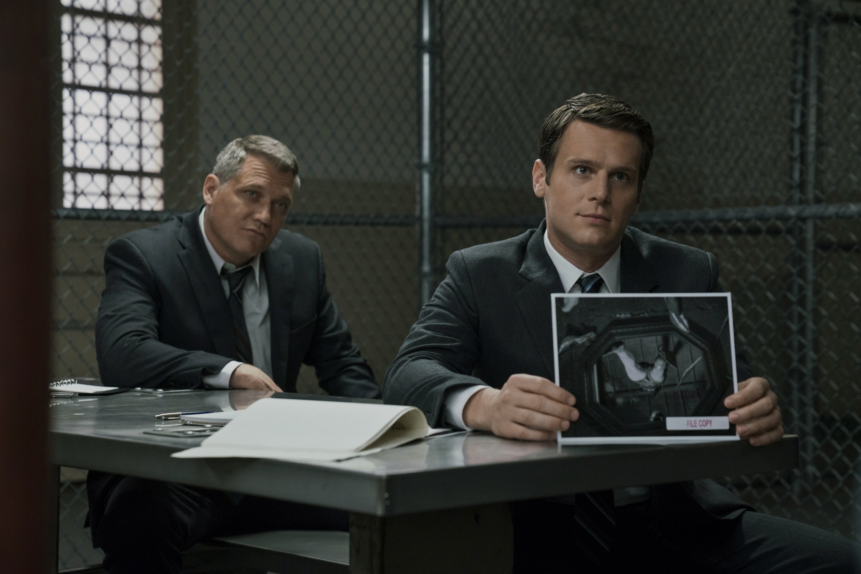 Two detectives on one side of an interrogation table show evidence to someone out of frame.
