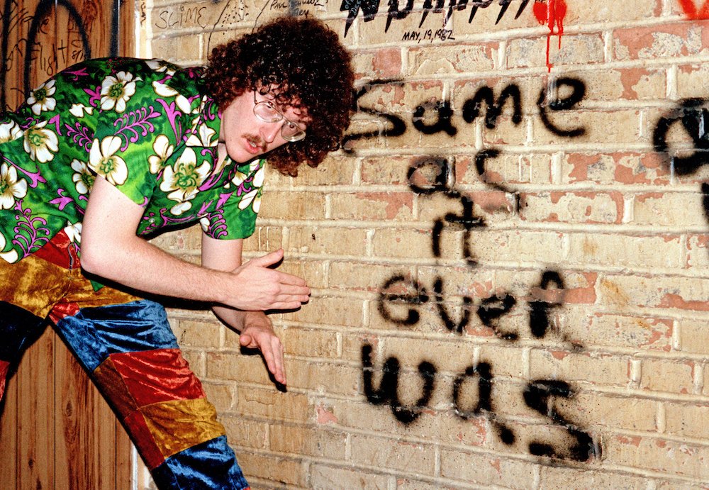 Weird Al Yankovic in a green Hawaiian shirt and stripy pants doing a robot pose in front of a brick wall. Graffiti on the wall says 