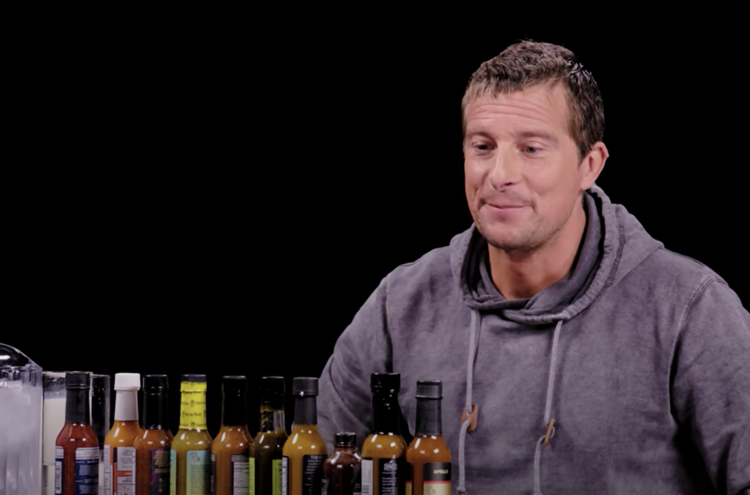Bear Grylls with a bunch of spicy sauces in front of him.