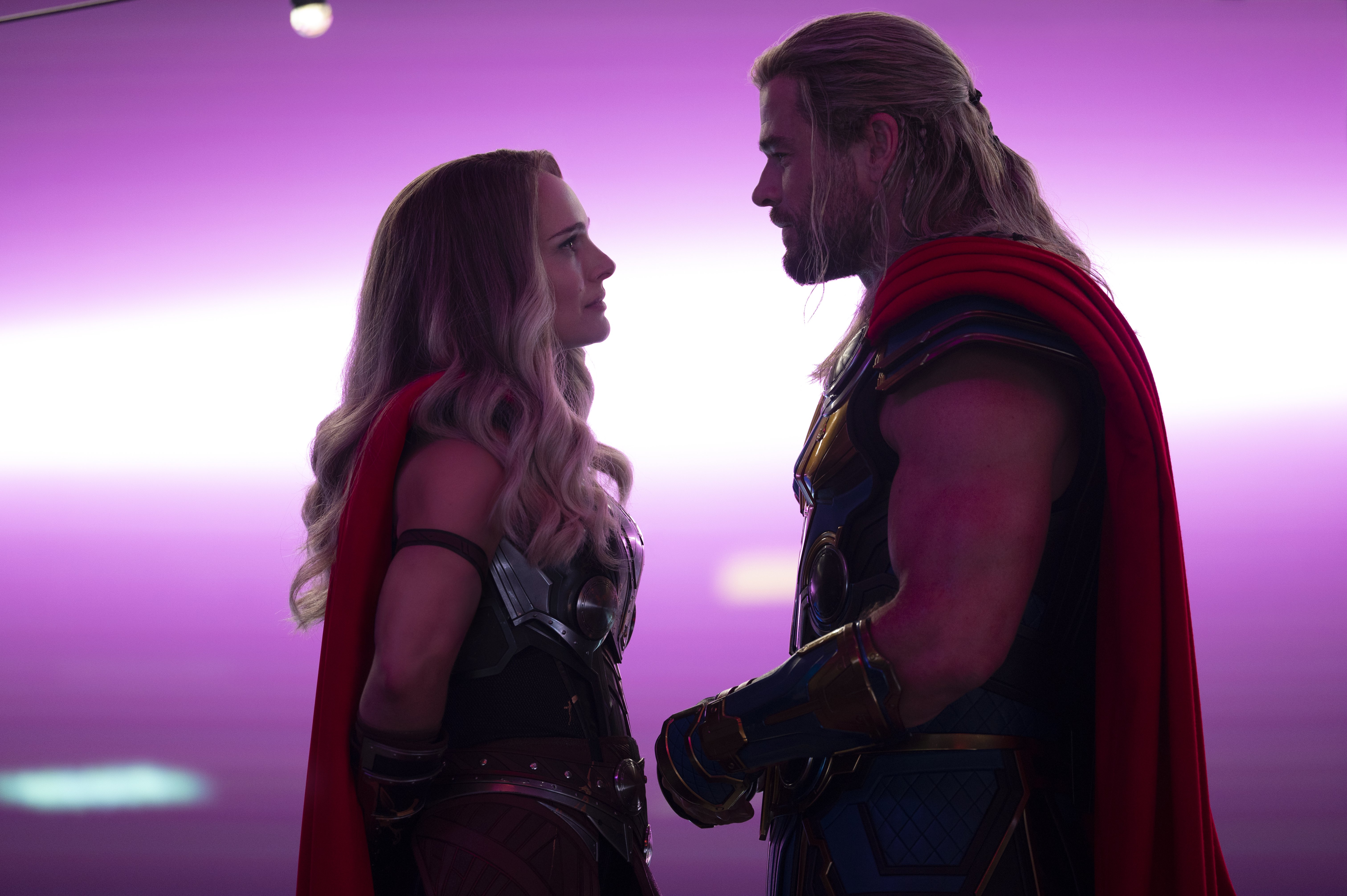 A man and woman in Nordic armor and red capes look at each other while purple light glows behind them.