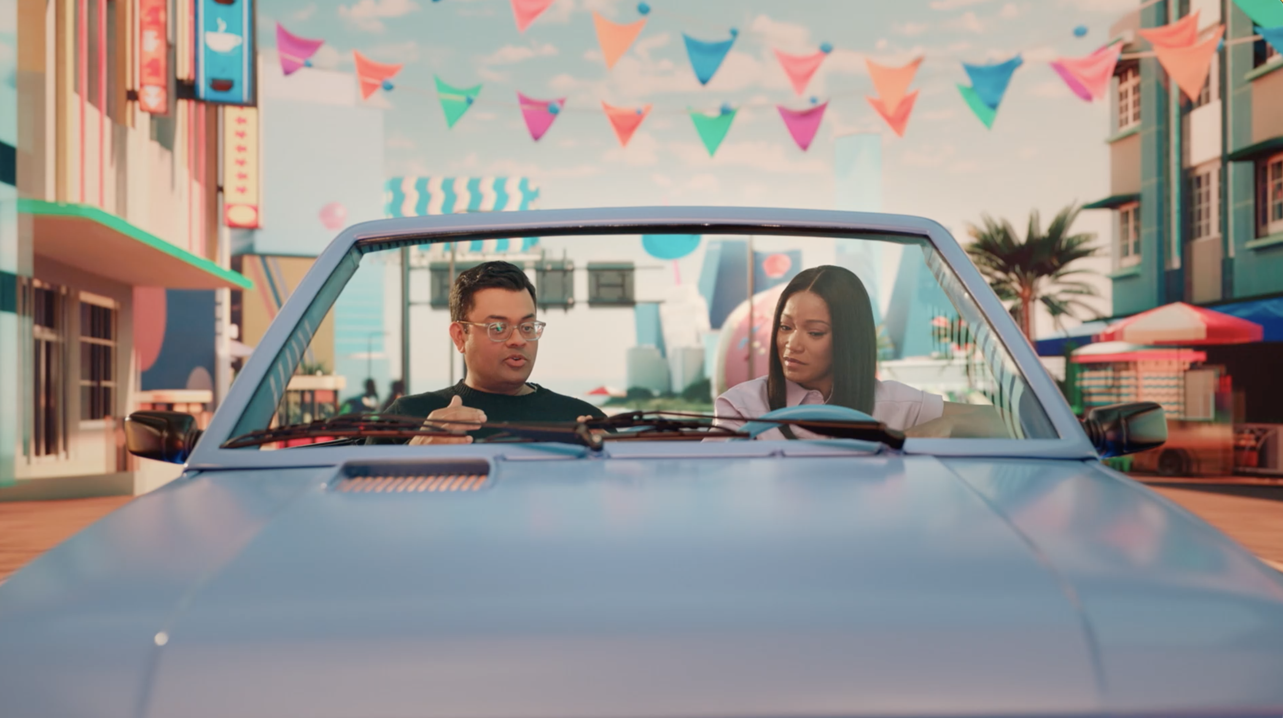 Keke Palmer and Vishal Shah sit in the driver and front seat of a blue open-top convertible driving down a Miami-inspired street.