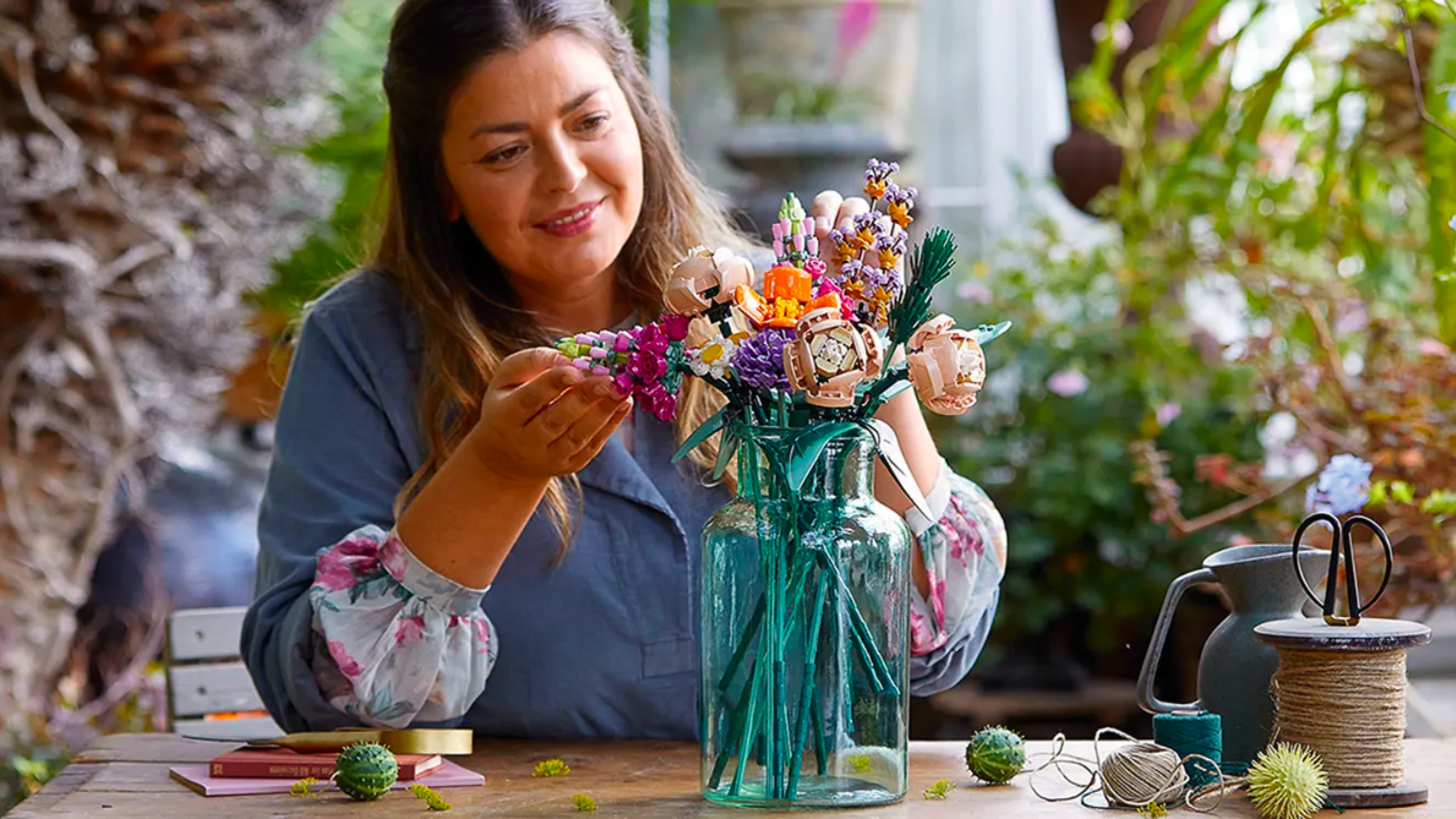 a woman in a blue shift with floral sleeves working on the lego flower bouquet kit at a crafting table